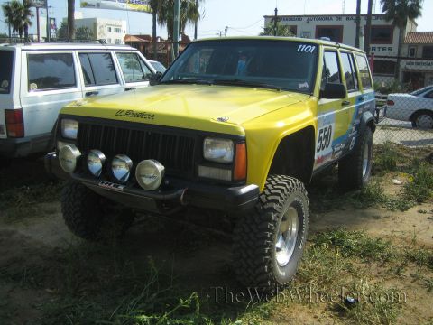 1990 Ford Bronco - yellow Jeep Cherokee race vehicle numbered 550 with four front driving lights and big tires sits on a dirt lot by palm trees.