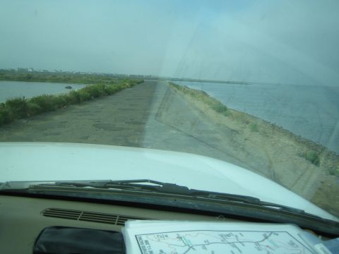1990 Ford Bronco - Seen through a windshield, a rough dirt causeway runs between two bodies of water with a folded map resting on the dashboard.