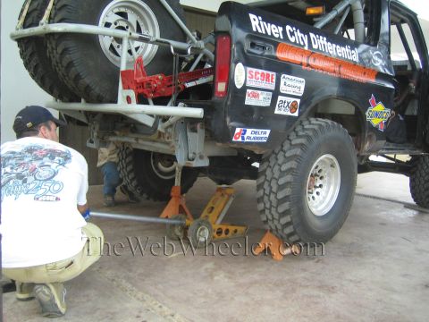 1990 Ford Bronco - kneeling man works under a lifted black off-road Ford Bronco marked River City Differential, raised on a floor jack in a shop.