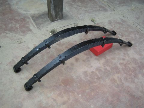 1990 Ford Bronco - Two heavy black multi-leaf truck suspension springs rest on a concrete floor next to a small red box.