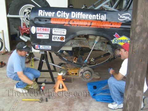 1990 Ford Bronco - Two kneeling men work on the bare chassis of a black West Coast Broncos race vehicle supported on jack stands in a shop.