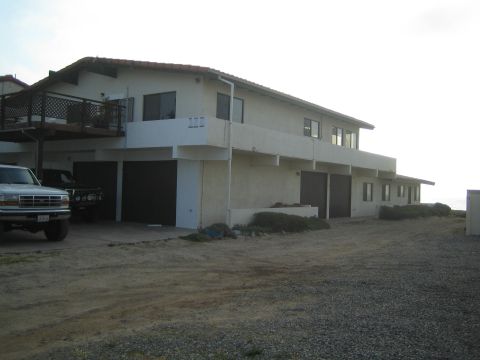1990 Ford Bronco - two-story white stucco building with an upper deck sits on a gravel lot, a lifted white pickup parked beneath one end.