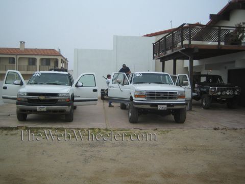 1990 Ford Bronco - white Chevrolet pickup and a white Ford crew-cab pickup park on a dirt lot beside a stucco building, a dark Bronco at right.