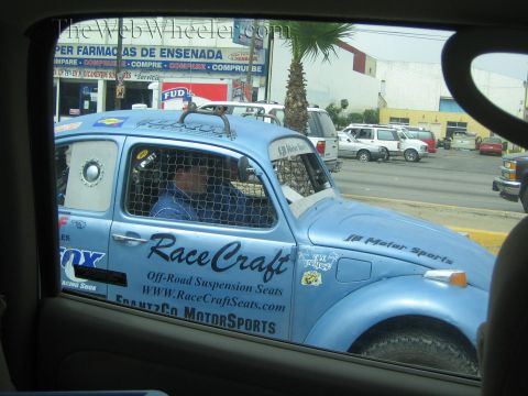 1990 Ford Bronco - blue Baja-style Volkswagen Beetle race car lettered RaceCraft drives a Mexican street, viewed through a vehicle window.