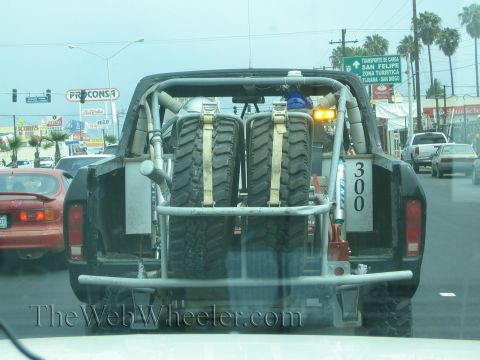 1990 Ford Bronco - rear of a black off-road prerunner truck carrying two mounted spare tires sits in traffic on a Mexican street.