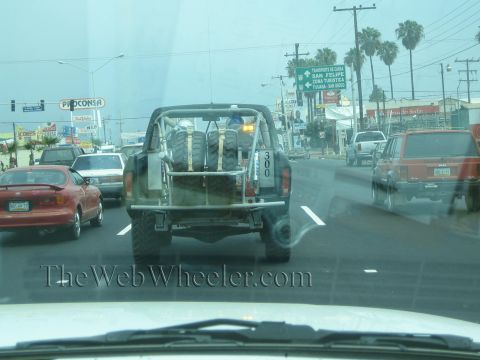 1990 Ford Bronco - black prerunner truck with bed-mounted spare tires drives ahead in busy traffic, seen through a rain-flecked windshield.