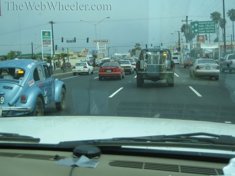 1990 Ford Bronco - blue VW Beetle race car and a gray prerunner truck travel a busy Mexican boulevard, viewed through a car windshield.