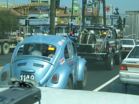 1990 Ford Bronco - blue Baja Beetle numbered 1149 follows a truck towing a trailered off-road race buggy through a Mexican street with shops.
