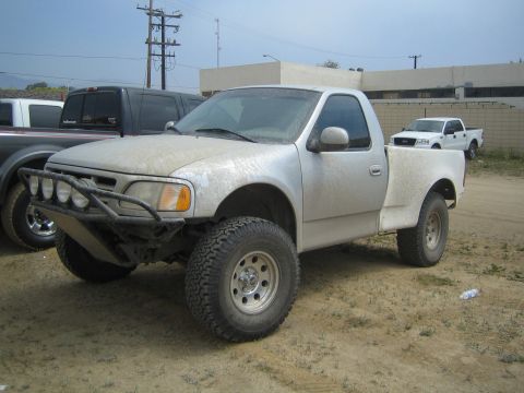 1990 Ford Bronco - dusty white Ford regular-cab pickup fitted with a tubular prerunner bumper and light bar sits on a dirt lot.