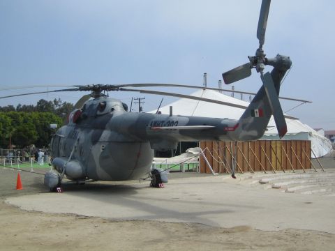 1990 Ford Bronco - large gray-and-blue camouflaged military transport helicopter sits parked on a dirt lot, a Mexican flag on its tail.