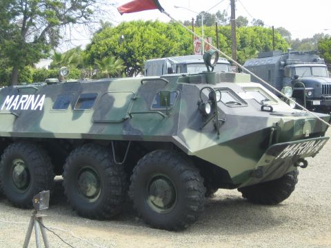 1990 Ford Bronco - green-camouflage six-wheeled armored personnel carrier marked MARINA is displayed outdoors among trees and other vehicles.