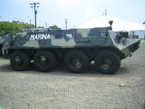 1990 Ford Bronco - Side profile of a green-camouflage eight-wheeled armored personnel carrier marked MARINA parked on a gravel display lot.