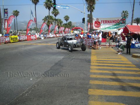 1990 Ford Bronco - open-frame off-road race car waits at a street start line flanked by Tecate banners, sponsor flags, palms and spectators.