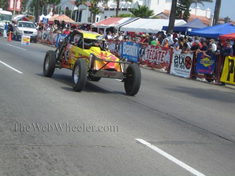 1990 Ford Bronco - yellow-and-red open-frame off-road buggy races down a street course lined with sponsor banners and a packed crowd.