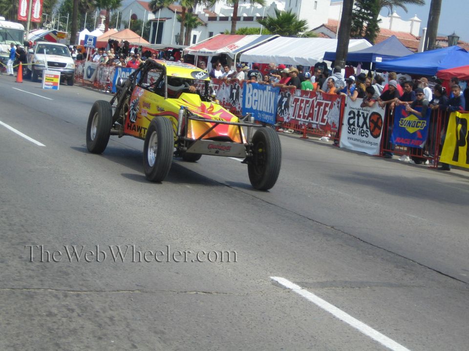 Yellow and red open-frame off-road buggy racing on a street course lined with crowds and banners