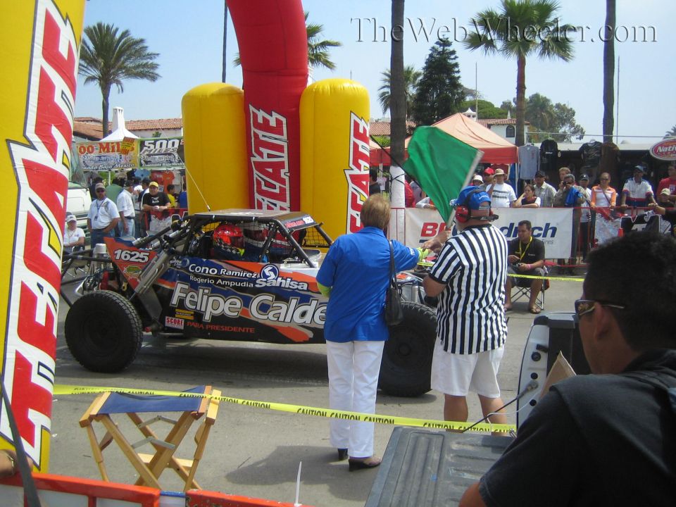 Red number-1625 off-road race truck at a Tecate arch start with officials and crowd