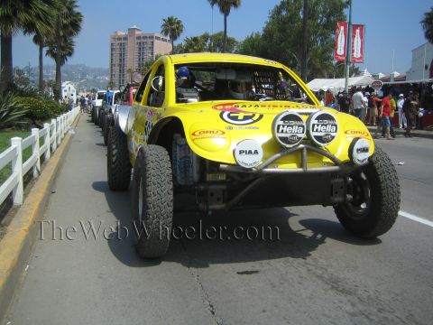 1990 Ford Bronco - yellow Class 5 Baja Bug race car with Hella and PIAA lights sits on a palm-lined street during a desert race contingency.