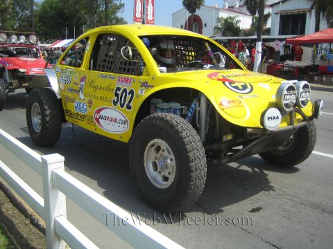 1990 Ford Bronco - Yellow off-road race Beetle marked 502 covered in sponsor decals including BFGoodrich and Hella, staged near event tents.