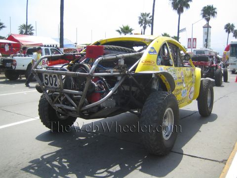 1990 Ford Bronco - Rear of yellow off-road race buggy 502 exposing its rear engine, tube frame and spare tire among palms and trucks.