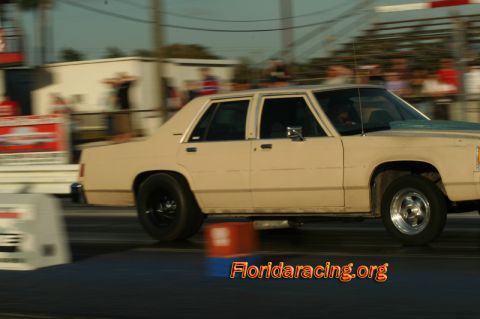 1985 Ford Crown Victoria LTD - A tan sedan is captured in motion on a drag strip, with spectators visible in the background.