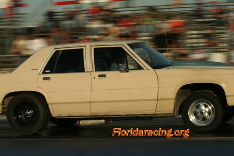 1985 Ford Crown Victoria LTD - A tan sedan with a dark hood is captured in motion at a drag strip, with spectators blurred in the background.