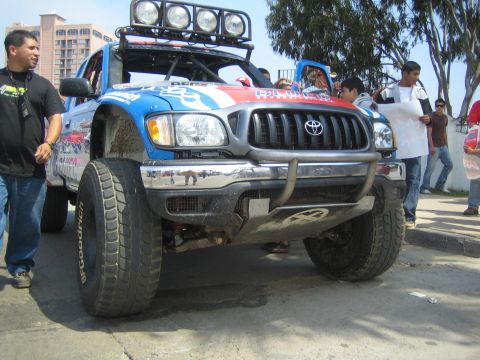 1990 Ford Bronco - muddy blue and red Toyota Tacoma prerunner truck with a roof light bar and tube bumper draws onlookers on a street.