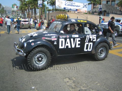 1990 Ford Bronco - Black off-road race Beetle marked DAVE CO 1145 with roof lights idles on a street below a Felipe Calderon banner.