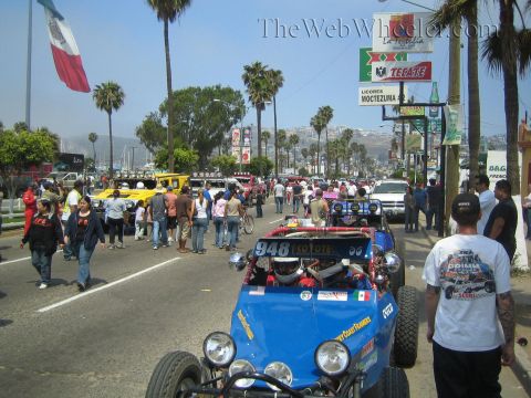 1990 Ford Bronco - Blue single-seat off-road buggy 948 rolls through a crowd of spectators on an avenue with a Mexican flag and Tecate signs.