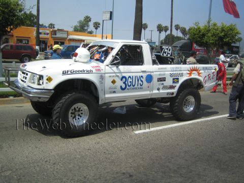 1990 Ford Bronco - White off-road race truck with 3Guys Autowrecking and BFGoodrich decals staged by spectators on a palm-lined street.