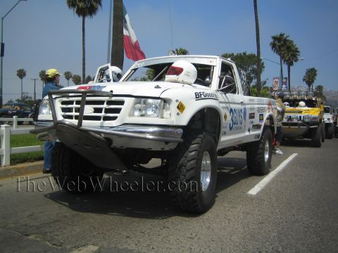 1990 Ford Bronco - White Ford race truck with chrome tube bumper and BFGoodrich livery, helmeted crew nearby and a Mexican flag behind.