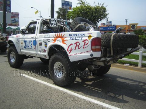 1990 Ford Bronco - Rear of white off-road truck 759 lettered GRP Horizon Realty carrying mounted spare tires on a street with shops behind.