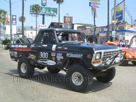1990 Ford Bronco - Black off-road Bronco 300 with Deaver Suspension livery drives past colorful storefront signs including Licores King.