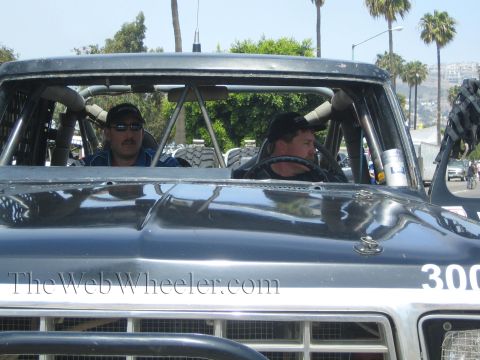 1990 Ford Bronco - Two men sit in the caged cab of a dark off-road race truck numbered 300, seen across its hood on a sunny street.