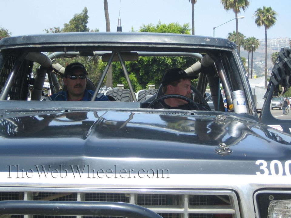 Two men seated in the cab of a dark off-road race truck viewed over the hood
