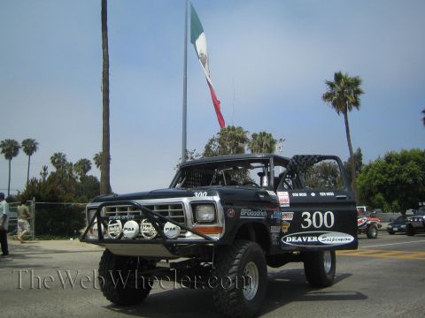 1990 Ford Bronco - Black off-road Bronco 300 with Deaver Suspension and roof lights staged beneath a tall Mexican flag among palms.