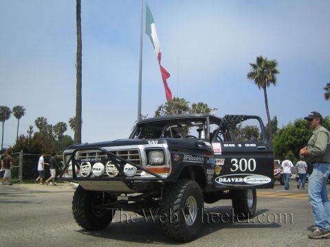 1990 Ford Bronco - Black off-road Bronco 300 covered in sponsor decals sits on a street as a man passes and a Mexican flag flies behind.