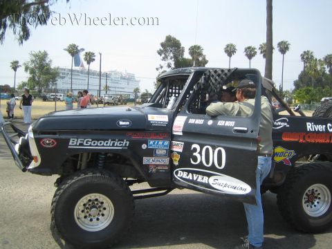 1990 Ford Bronco - Black off-road Bronco 300 with crew at the door is staged on grass with a large cruise ship and palms in the background.
