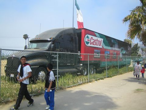 1990 Ford Bronco - black semi tractor with a red Castrol-branded trailer sits behind a chain-link fence as two young men walk past.