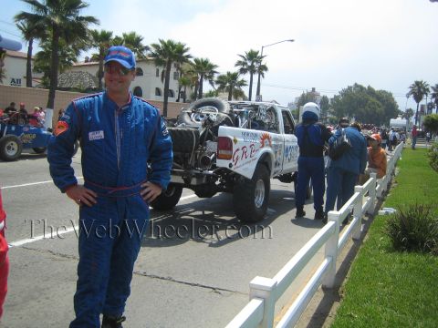 1990 Ford Bronco - man in a blue driving suit poses on a palm-lined street next to a white GRP off-road race truck and crew.
