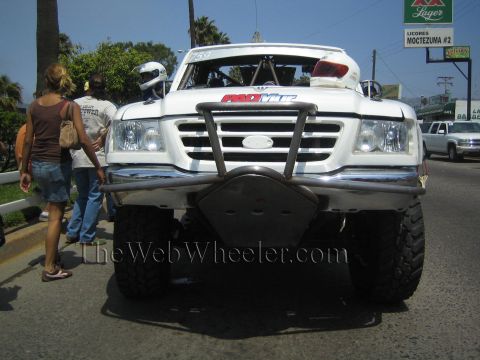 1990 Ford Bronco - White off-road race truck 759 with Pro Comp livery and a helmeted driver faces the camera as pedestrians pass on a street.