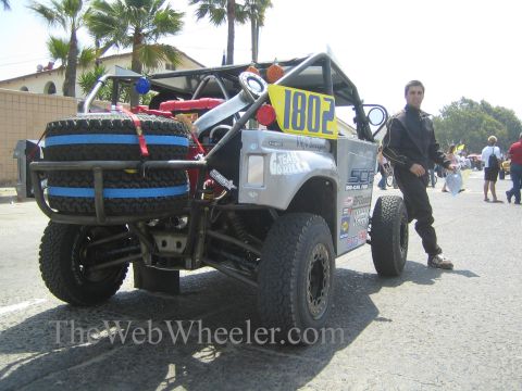 1990 Ford Bronco - Silver SCORE-class tube-frame race truck 1802 with stacked spare tires is staged on a street as a man stands at its side.