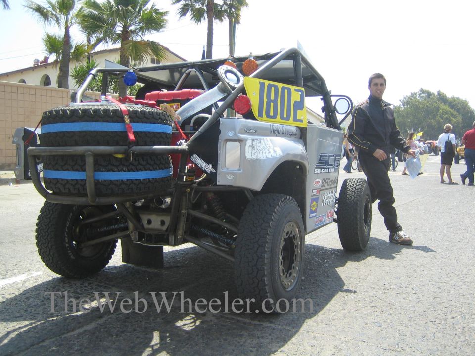 Silver tube-frame off-road race truck 1802 with spare tires and a man standing beside
