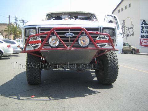 1990 Ford Bronco - White Ford off-road race truck with a red tube bumper and four round driving lights faces the camera on a town street.