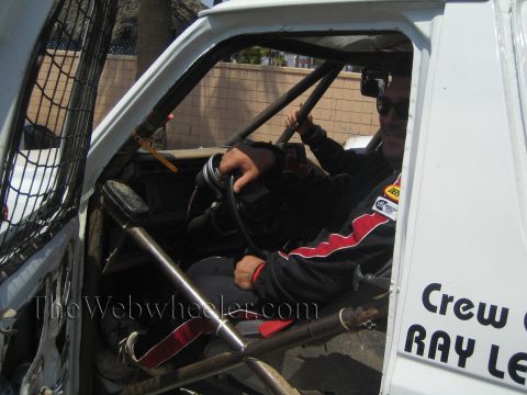 1990 Ford Bronco - driver in a red-and-black race suit sits behind the wheel inside the caged cab of a white off-road race truck.