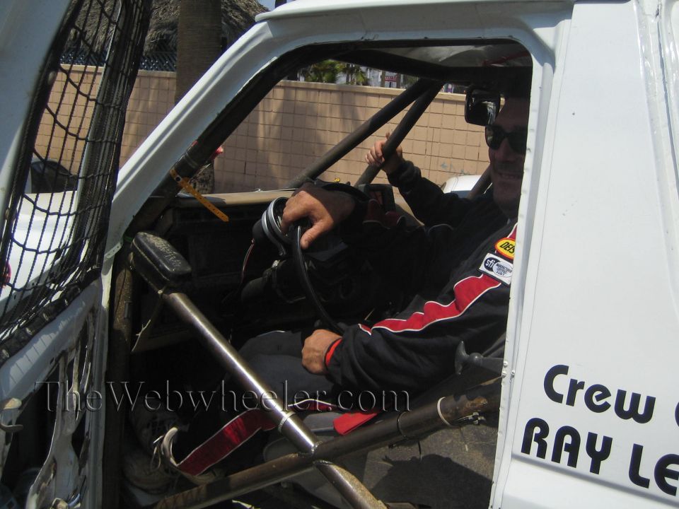 Driver in red and black racing suit seated inside white off-road race truck cab with roll cage