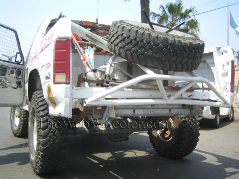 1990 Ford Bronco - Rear of a white off-road race truck showing a tubular rear bumper, mounted spare tire and oversized mud-terrain tires.