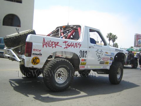 1990 Ford Bronco - white lifted Ford Bronco race truck marked number 302 and 'Anger Issues' sits on a city street covered in sponsor decals.