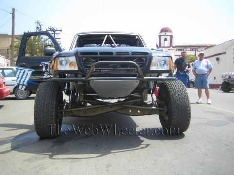 1990 Ford Bronco - Head-on view of a dark off-road race truck with a tubular front bumper and long-travel suspension as two men stand nearby.