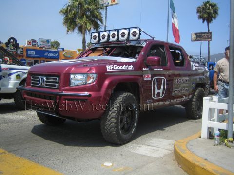 1990 Ford Bronco - maroon Honda Ridgeline off-road race truck with a roof light bar and BFGoodrich livery parked near palm trees and a Mexican flag.
