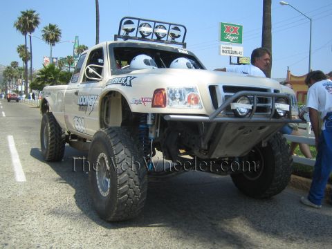 1990 Ford Bronco - tan Ford Ranger prerunner race truck with roof lights and helmeted occupants moves down a palm-lined street past spectators.